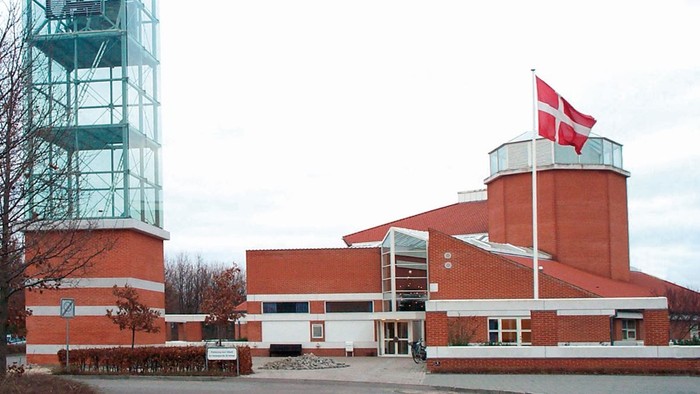 Bygningen har en stor glasstruktur og dansk flag.