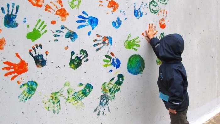 Ein Kind macht einen farbigen Handabdruck an eine Wand. Es sind schon viele bunte Handabdrücke auf der Wand