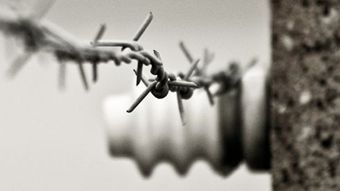 Eine schwarz-weiße Nahaufnahme zeigt ein Stück Stacheldraht, das sich diagonal durch das Bild zieht. Der Draht ist scharf fokussiert, einzelne Widerhaken sind deutlich zu erkennen. Im unscharfen Hintergrund sind schemenhaft eine Mauer oder ein Pfeiler sow