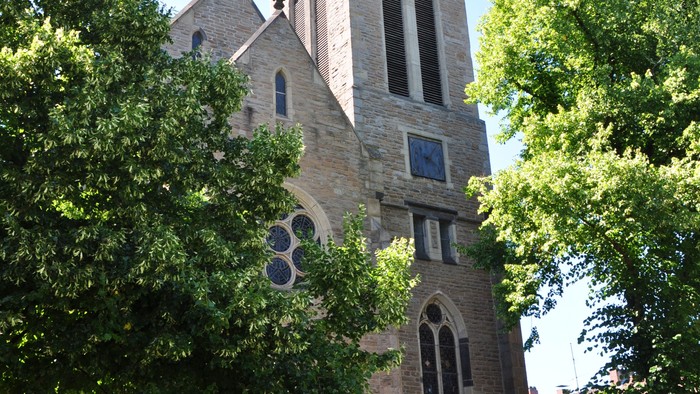 Historische Steinkirche mit hohem Turm und Bogenfenstern, umgeben von üppigen grünen Bäumen.