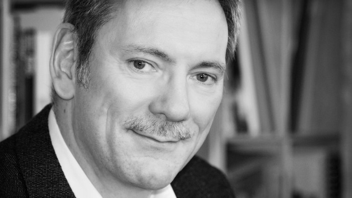 En mand med overskæg iført jakkesæt og hvid skjorte, der poserer i et professionelt sort-hvidt portræt.