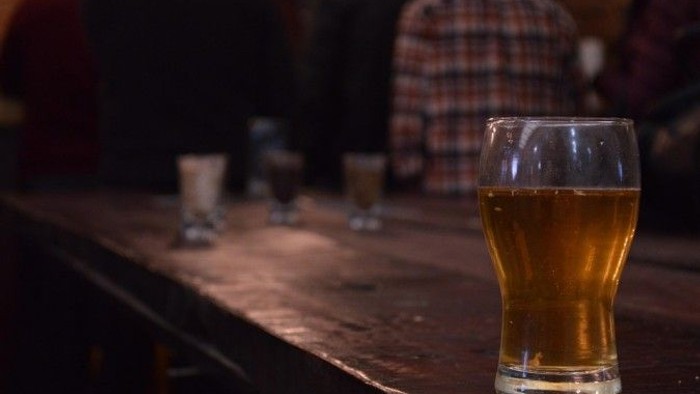 Glas mit hellem Bier auf einem dunklen Holztisch in einer gemütlichen Bar- oder Kneipenatmosphäre, weitere Gläser unscharf im Hintergrund.