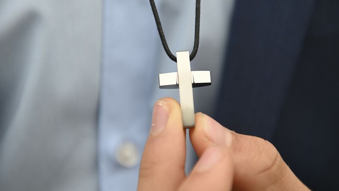 Eine Hand hält ein silbernes Kreuz an einer schwarzen Kette vor einem blauen Hemd.
