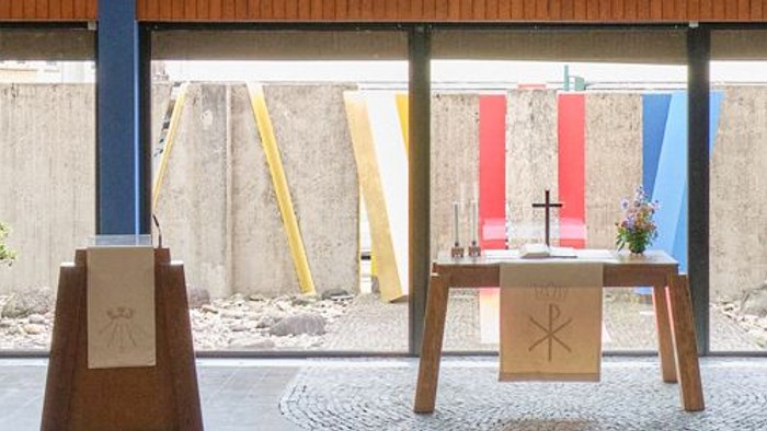 Minimalistische Outdoor-Kapellen-Einrichtung mit hölzernen Podesten, religiösen Symbolen und farbenfrohen geometrischen Wänden im Hintergrund.