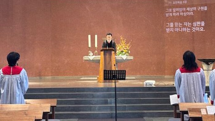 Ein Gottesdienst ist im Gange, bei dem ein Redner an einem Pult in einer Kirche steht, flankiert von zwei Personen in Roben.