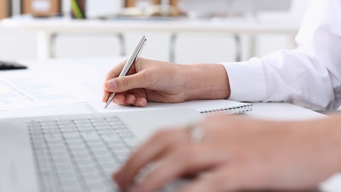 En person skriver med pen på et papir og bruger samtidig en laptop.