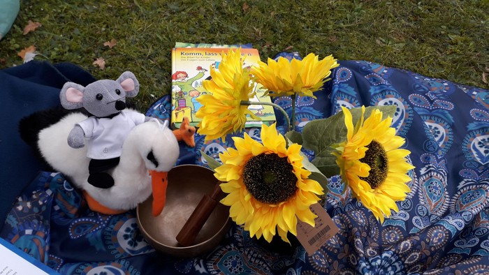 Ein Stofftier, Sonnenblumen und ein Buch liegen auf einem blauen Tuch im Gras.
