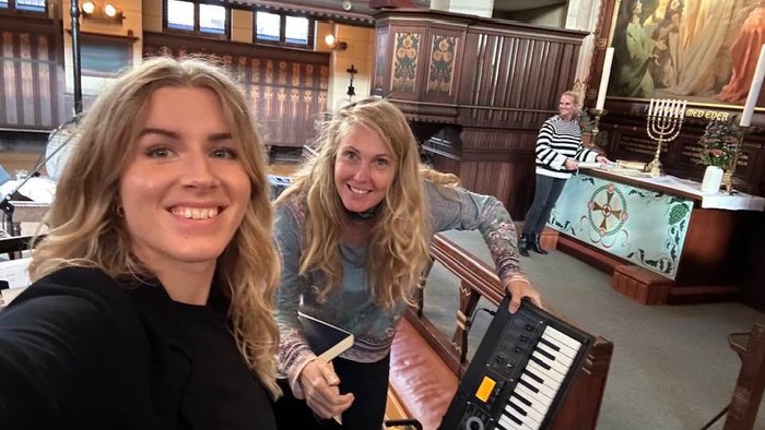 To kvinder smiler, mens de poser med et keyboardorgel i en prægtig indendørs omgivelse.