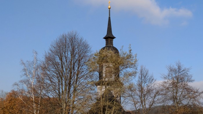 Ein hoher Kirchturm ragt hinter kahlen Bäumen in den klaren blauen Himmel.