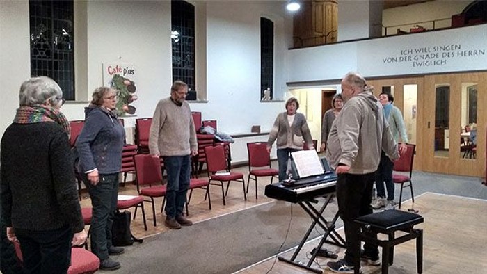 Eine Gruppe von Menschen probt in einem Raum mit Klavier und Stühlen.