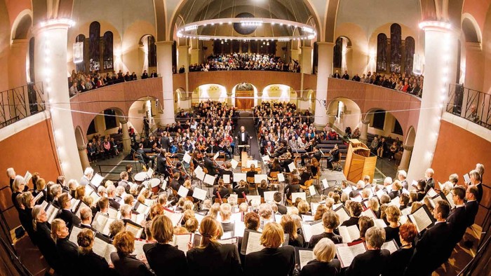 Ein großer Konzertsaal, gefüllt mit einem Publikum und einem Orchester, das auf der Bühne spielt.