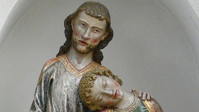 Holzstatue, die einen bärtigen Mann darstellt, der ein schlafendes Kind in traditionellen Gewändern zärtlich hält.