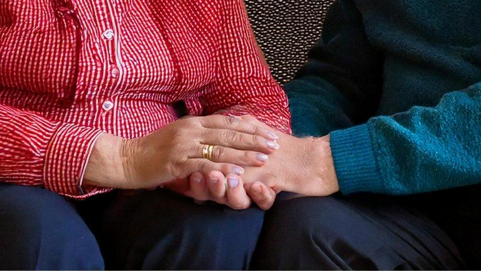 Zwei Personen, die sich an den Händen halten: eine trägt ein rotes kariertes Hemd, die andere einen blauen Pullover.