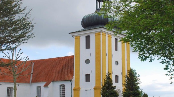 En hvid og gul kirke med sort tårn og rødt tag, omgivet af træer og en stenmur.