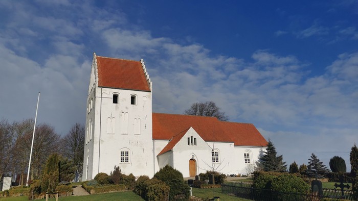 Hvid kirke med rødt tag og tårn, omgivet af græs og buske