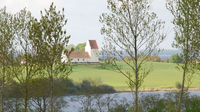 En idyllisk landskab med en sø, grønne felter og et hvidt hus med røde tag.