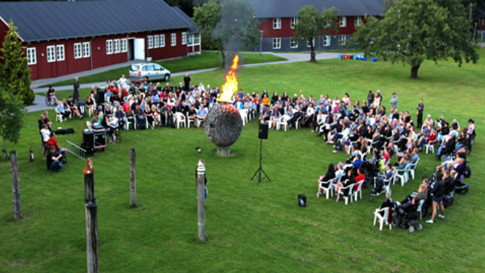 Udendørs sammenkomst med en stor folkemængde siddende på stole og iagttagende et bål og en optræden på en græsklædt mark.
