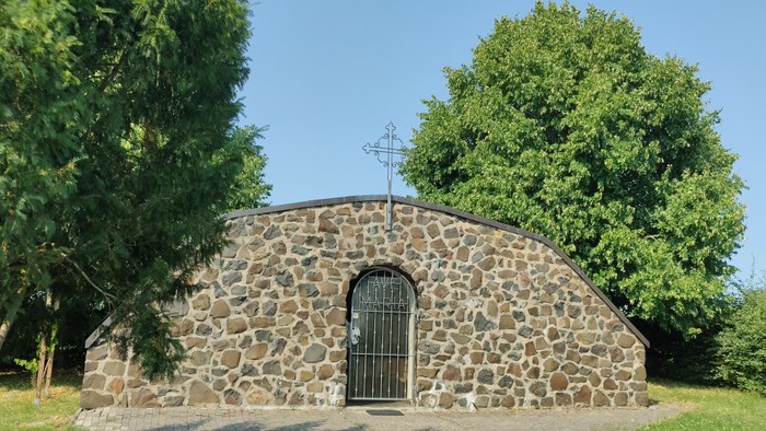 Kleine steinerne Kapelle mit Kreuz auf dem Dach, umgeben von Bäumen und Gras