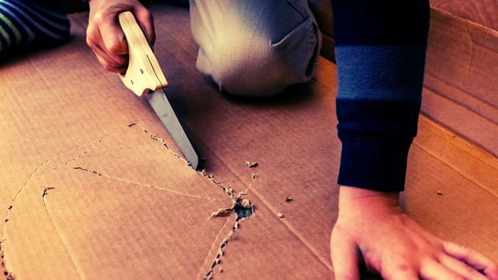 Eine Person schneidet vorsichtig Pappe mit einem Cuttermesser entlang einer vorgezeichneten Linie.