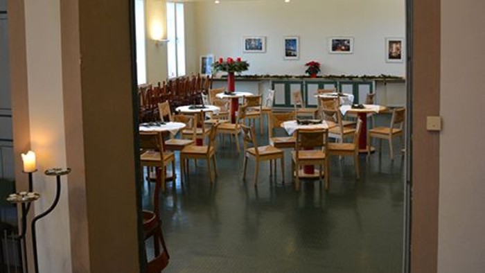 Der Blick fällt durch eine offene Tür in einen hellen, ruhigen Raum, der als Café- oder Veranstaltungsbereich genutzt wird. Mehrere kleine runde Tische sind gleichmäßig im Raum verteilt, jeweils mit Stühlen aus hellem Holz. Auf den Tischen liegen Tischset