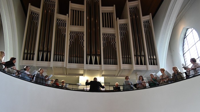 Ein großes Orgelkonzert mit einem Publikum, das sich auf einem Balkon in einem geräumigen, eleganten Konzertsaal versammelt hat.