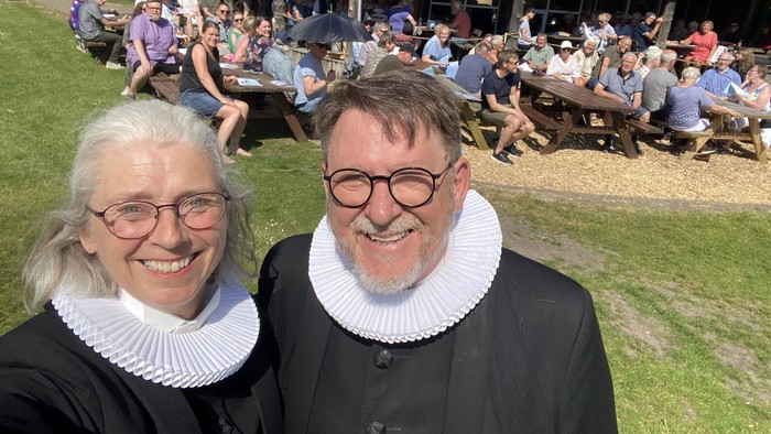 En mand og en kvinde i historiske kragekraver står foran en folkemængde ved en træbygning.