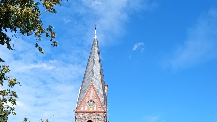 Kirchengebäude mit hohem Turm und spitzem Dach, umgeben von Bäumen und Büschen