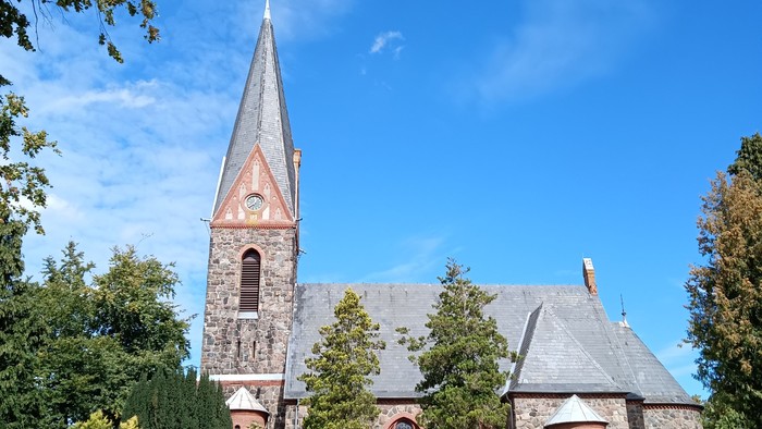 Kirchengebäude mit hohem Turm und spitzem Dach, umgeben von Bäumen und Büschen