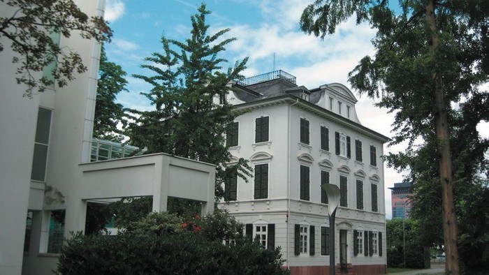 Eleganter historischer Bau, umgeben von modernen Gebäuden und üppigem Grün unter klarem Himmel.