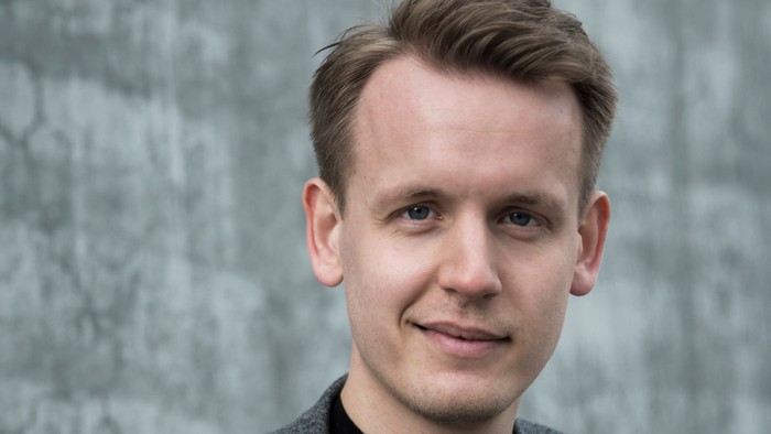 En smilende mand med kort, brunt hår iført en mørk skjorte og en grå blazer foran en tekstureret baggrund.