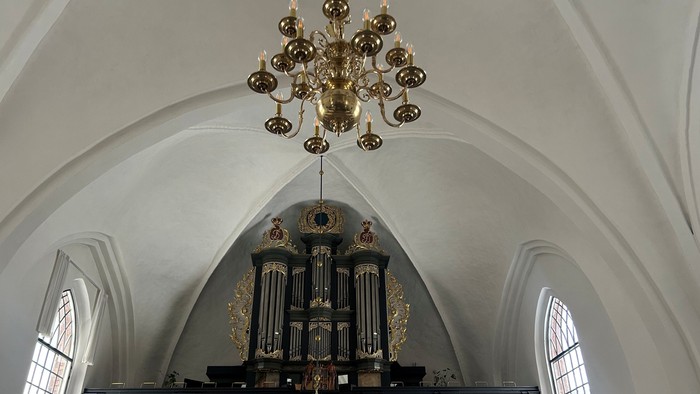 Elegant kirkeinteriør med et prægtigt pibeorgel og en stor hængende lysekrone under hvælvede lofter.