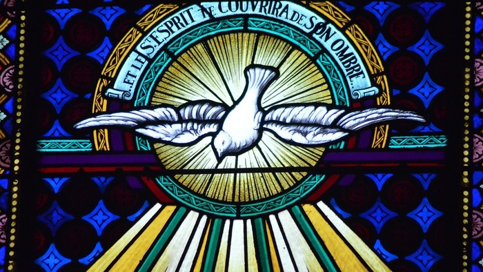 Das Bild zeigt ein buntes Kirchenfenster mit einer Taube und lateinischem Text.