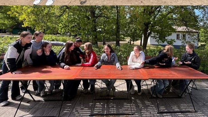 Große Gruppe von Menschen sitzt an langen Tischen im Freien, scheint ein Treffen oder ein Picknick zu sein.