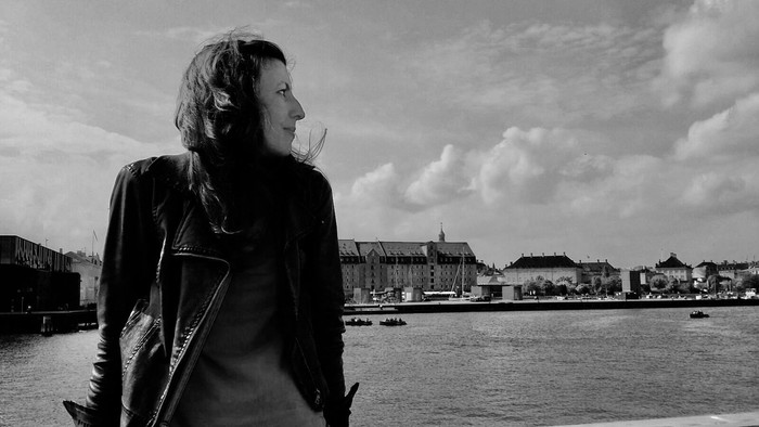 En eftertænksom person står ved kajen og kigger ud over et malerisk bylandskab og vandet.