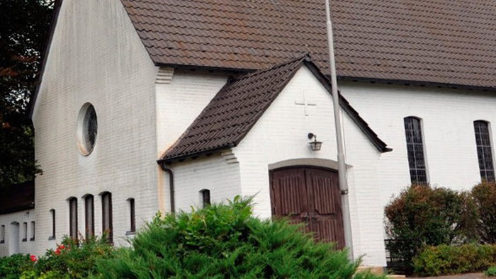 Weiße Kirche mit hohem Turm und braunem Dach, umgeben von Bäumen und Büschen