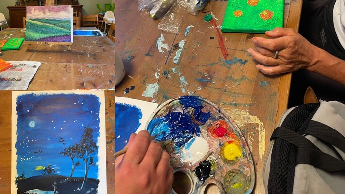 Kreativer Arbeitsbereich mit lebendigen Gemälden, Kunstmaterialien und Händen, die Farbe auf einer Palette mischen.