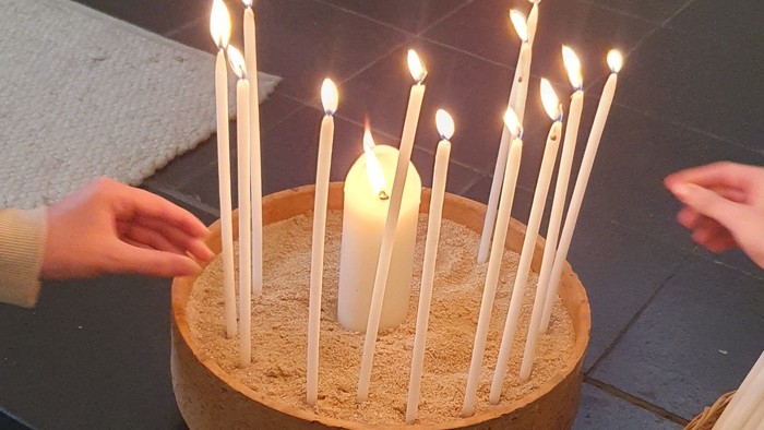 Ein runder, mit Sand gefüllter Behälter hält zahlreiche brennende Kerzen, während Hände in der Nähe möglicherweise Vorbereitungen für eine Feier treffen.