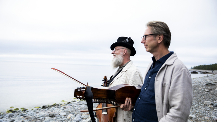 To mænd spiller strengeinstrumenter ved kysten på en stenet strand.