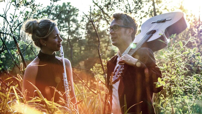 To personer på en solbeskinnet mark, hvor den ene holder en guitar, mens de fører en samtale omgivet af højt græs.