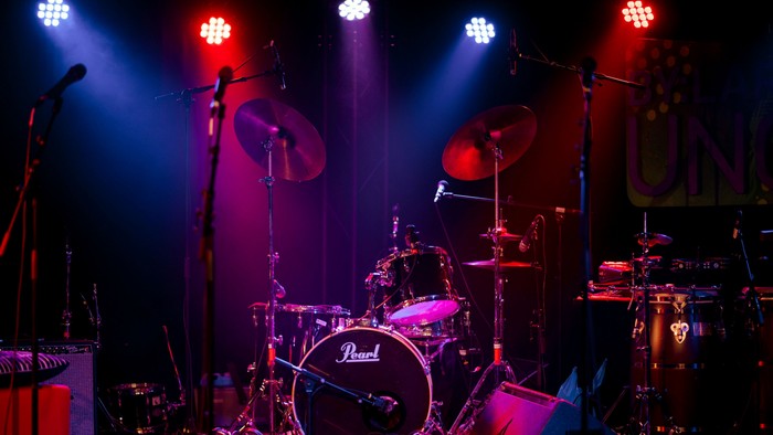 Das Bild zeigt ein Schlagzeug und Mikrophone in einem dunklen Raum mit bunter Beleuchtung.