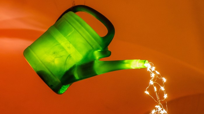 Eine leuchtend grüne Gießkanne, die Lichterketten auf einen warmen roten Hintergrund gießt.