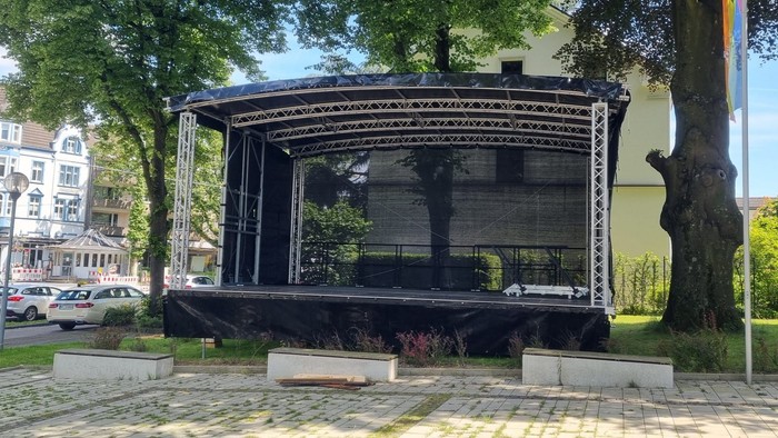 Ein offener Pavillon mit filigranem Design steht auf einem Kopfsteinpflasterplatz, umgeben von Bäumen und Gebäuden.