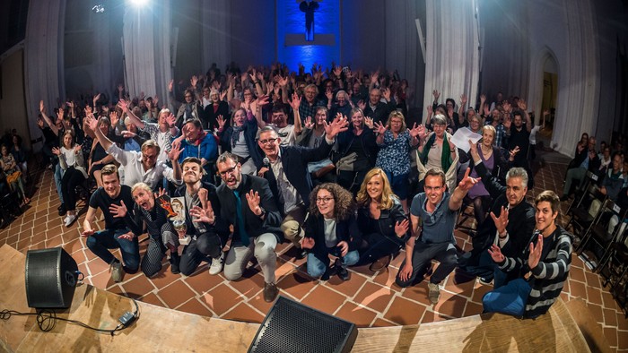 Große Gruppe von Menschen posiert in einem Raum mit hellen Lichtern und Vorhängen