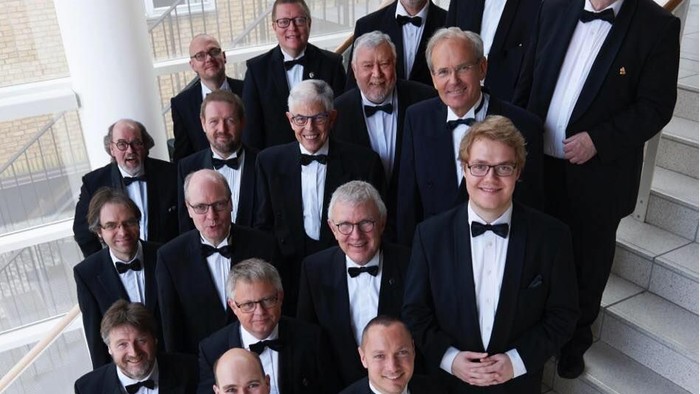 Gruppe af mænd i sorte smoking, der poser sammen på en stor indendørs trappe.