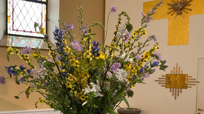 En levende buket i en metallisk vase står på et bord ved siden af et roligt kirkeinteriør med farvet glas.