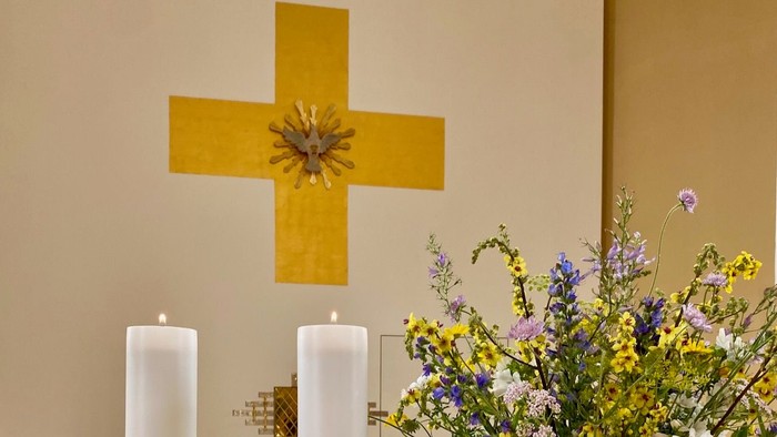 En gul korsformet vegn med blomster i midten, to lysede lysstaker og en blomsterarrangement på alteret