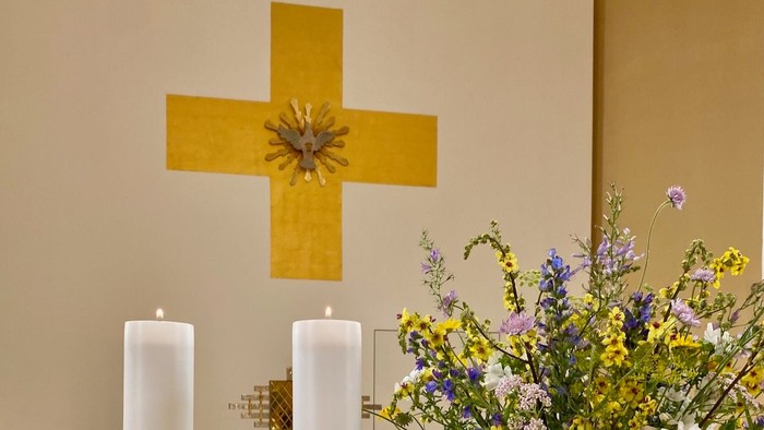 En gul korsformet vegn med blomster i midten, to lysede lysstaker og en blomsterarrangement på alteret