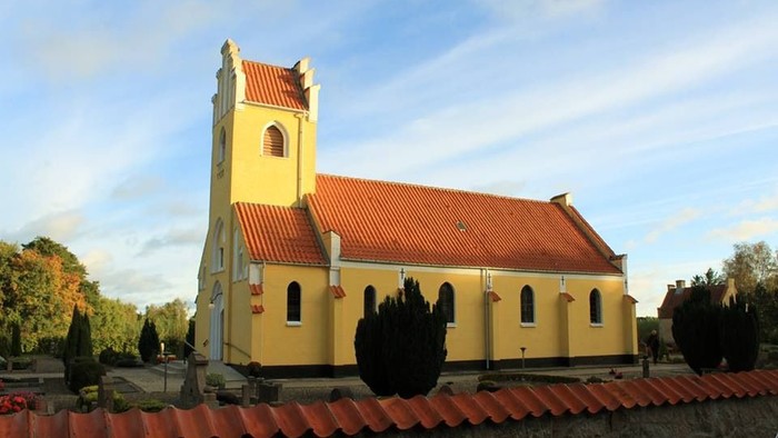 Gul kirke med rødt tag og tårn, omgivet af træer og mur.