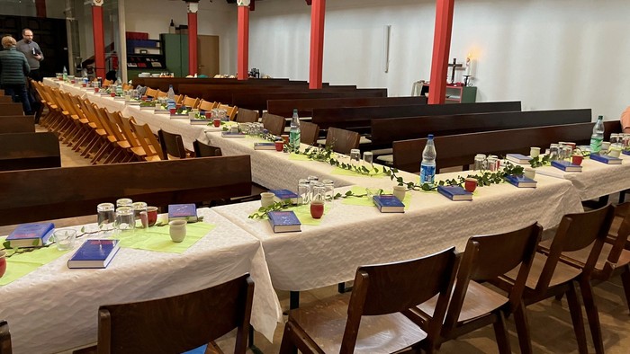 Ein geräumiger Saal mit langen Tischen, die für eine formelle Veranstaltung gedeckt sind, geschmückt mit Grünpflanzen und Wasserflaschen.