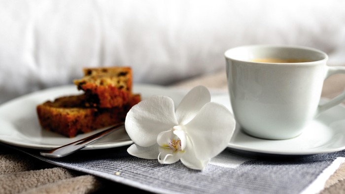 Eine gemütliche Platte mit einem Stück Kuchen, einer weißen Orchidee und einer Tasse Kaffee auf einem weichen Tuch.